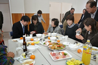 刺身や惣菜などが並ぶテーブルを囲んだ人々が、お酒をついだり、笑顔で談笑しながら楽しんでいる様子の写真