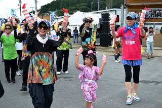 浴衣姿の幼い女の子とカラフルな服装の大人たちが並び、全員が手に赤と白の鳴子を持って楽しそうに演舞している写真