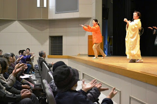 舞台の上で黄色の着物を着た女性やスタッフが手のひらを上にし両手を前に出している動きをし、前列に座っている参加者が楽しそうに一緒に手を動かしている様子が写っている写真