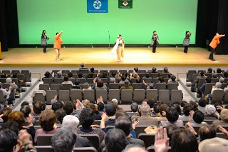 舞台に6名の人が並んで観客席に手をあげている様子を写した写真