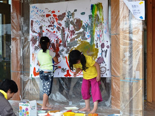 女の子2人で壁に貼られた大きな紙に絵の具を使って自由に絵を描いており、色とりどりの筆跡が紙いっぱいに広がっている様子が写っている写真