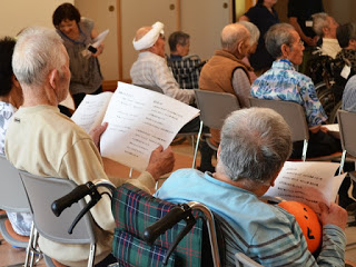 椅子に座った高齢者の方々が楽譜をもって一緒に歌を歌っている様子を後方から写した写真