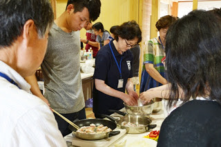 数人の男女がテーブルで協力しながら、フライパンで肉団子のような料理を焼いたり、ボウルで材料を混ぜたりしている様子の写真