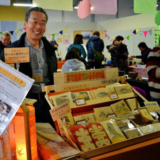 木製の商品棚に魚や木製キーホルダーなどの手づくり品を並べて笑顔で立っている男性の販売者と、その周囲で買い物をしている人々の様子が写っている写真