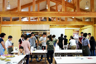 天井の高い木造の室内で、多くの参加者がテーブルの周りで料理の準備をしている写真