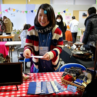 テーブルに布製品や小物が並び、「アイロンビーズ」と書かれた札が掲げられた手作り品販売ブースの前に、カラフルなセーターを着て紙を持った笑顔の女性が接客している様子を写した写真