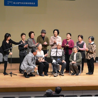 椅子に3名の方が座り後ろに7名の人が手拍子をしながら歌を歌い関係者2名がマイクをかたむけている舞台での写真