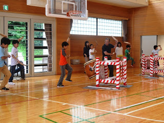 体育館の端で、複数の子どもたちが大太鼓の前に並び、指導者の合図で太鼓を叩く練習をしている写真