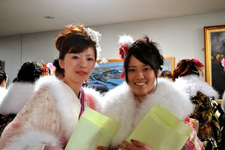 手には式の案内用紙が入った紙袋を持ち、振袖に白いファーを羽織った二人の若い女性が笑顔で並んでいる写真