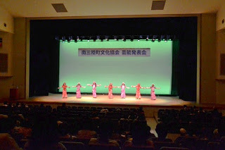 「南三陸町文化協会 芸能発表会」と書かれた横断幕が掲げられた明るいステージで、ピンクの衣装を着た女性たちが横一列になりフラダンスを踊っている様子を客席から写した写真
