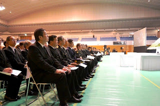 ベイサイドアリーナの広い屋内会場で、並べられた椅子に多くのスーツ姿の人々が整然と前を向いて座り、奥には観客席やステージがある式典の写真