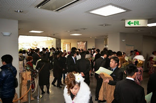 成人式の会場のロビーにスーツや振袖姿の多くの若者が集まり、案内用紙を手にして歩いたり話したりしている写真