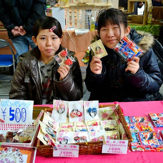 2人の子どもが販売ブースの席に座って商品を手に持ち、テーブルの上には値札が付いた手作り品が並んでいる様子を写した写真