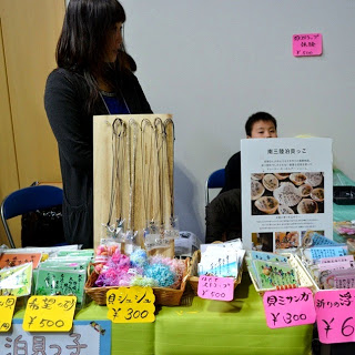 色とりどりの手作り雑貨や紙製品が並び、商品ごとにカラフルな価格札が貼られたテーブルの後ろで、女性と子どもが販売ブースに立っている様子を写した写真