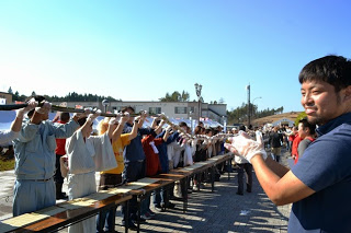 長テーブルに沿って並んだ手袋をつけた参加者が、一斉に巻き寿司を両手で持ち上げている写真
