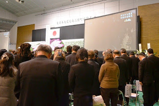 天井が高く広い室内会場で行われている式典で、多くの人々が前方を向いて立っており、正面には日本国旗と映像が映し出された大きなスクリーンがある写真