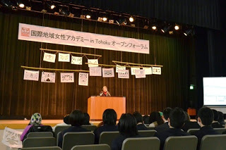ステージの背景には「国際地域女性アカデミー in Tohoku オープンフォーラム」と書かれた横断幕が掲げられ、その下には絵や文字が描かれた紙がいくつか吊るされ、ステージ上で講演をしている人物とそれを聴いている観客の様子を写した写真