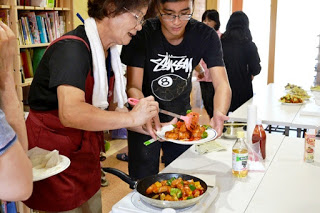 フライパンの中にはカラフルな野菜と肉の炒め物がある料理を、女性と男子学生が協力して皿に盛りつけている写真