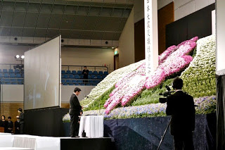 室内の大きな会場で行われている式典で、中央に白とピンクの花で飾られた大きな花壇があり、その前にテーブルと一人の男性が立っていて右側でカメラマンが撮影をしている写真