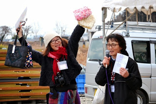 マイクを持って話している女性と、手作りの毛糸の帽子やバッグを手に笑顔で高く掲げている女性が、屋外のテントの下でマイクを持って話をしている様子が写っている写真。