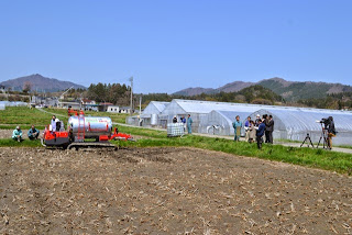 周囲にビニールハウスが並んでいる広い畑の中で液肥専用散布車が作業を行い、周囲で報道関係者や関係者が見守っている様子の写真