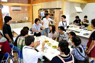 木の内装の部屋で、日本人と学生たちが一つのテーブルを囲み、料理を前に会話を楽しんでいる様子の写真