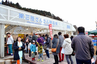 建物には大きな青い文字で「南三陸さんさん商店街」と書かれており、建物の入り口前には家族連れや観光客のような人々が買い物や見物に訪れている様子が写された写真