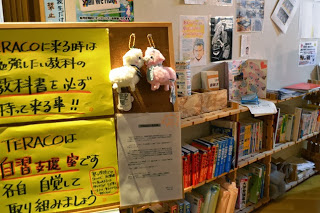 掲示物にかわいいぬいぐるみのキーホルダーと黄色い用紙に学習に関する貼り紙が張られ、本棚の上には受験に関する書物やたくさんの本が並んでいる室内の様子を写した写真