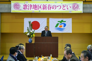 「歌津地区新春のつどい」と書かれた横断幕と国旗と町旗が掲げられた壇上で、スーツ姿の男性が演台に立って挨拶をしており、その前に着席した人々が話を聞いている写真