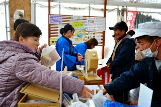 手前には、買い物をしている女性が店員と思われる男性から商品を受け取り、店の奥には黄色いガラポン抽選機が置かれており青い法被（はっぴ）を着たスタッフが複数人働いている 仮設テントのような構造の中を写した写真