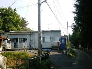 周囲に緑が生い茂る細い道沿いに立つ、自動車が停まっている簡易な白いプレハブの建物の写真