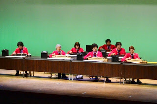 お揃いの赤い衣装を着た女性7人が舞台上の長机に座り、和楽器を演奏している写真