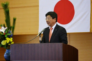 国旗と花の飾りのある演台に立ち、スーツとオレンジ色のネクタイを着た佐藤町長が挨拶を行っている写真