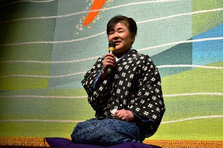 黒地に白い模様の着物を着た女性が舞台上で座布団に座り、マイクを持って語っている写真