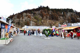 仮設店舗が左右両側に並び、中央部分にはハート型の緑の装飾や白い球体のオブジェが設置されその間を多くの人々が行き交う背後には山林が広がる丘が見えている屋外の仮設商店街を写した写真