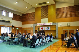 高い天井の会場で、複数のテーブルに分かれた参加者たちが座って式典を見守っている写真