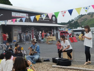 数人が屋外の広場でそれぞれ楽器を手に持ち、並んで演奏している様子が写っており、周囲には木々や建物が見える屋外演奏の写真