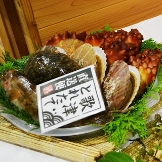 緑の葉で飾られた皿の上に貝類とウニが美しく盛り付けられており、中央には日本語で書かれた札が立てられている海産物の写真