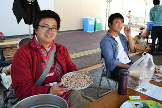 赤い上着を着た眼鏡をかけた男性が、小さな豆がたくさん入った金属皿を手に持って笑顔で見せている写真