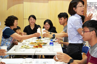 料理の並んだテーブルで、子供と女性と学生たちが紙コップを手に持ち、笑顔で乾杯をしている写真