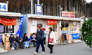 プレハブ型の小さな店舗が横並びになっている仮設商店街にはそれぞれに看板やのぼり旗が掲げられ、左側のベンチには数人の高齢者が座って談笑し手前では、防寒具を着た男女が歩いている様子を写した写真