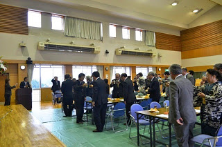 体育館のような広い会場でスーツ姿の人々が立ち上がり、テーブルを囲んで歓談しながら飲み物を手にし乾杯を行っている写真