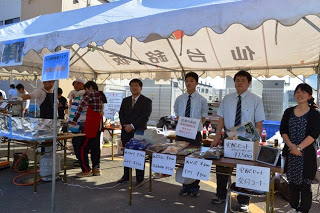 宅配セット受付コーナーと書かれたテントの下で、スーツ姿の男性3人と私服の男女が横一列に並んで立っている写真