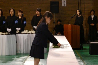 黒い服を着た参列者が見守る中、制服姿の少女が白いテーブルに花を手向けている献花の場面の写真