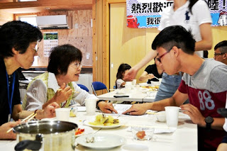 長テーブルを囲んで完成した料理を食べながら、年配の女性と男子学生が笑顔で会話している写真