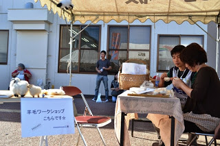 テントの下で「羊毛ワークショップ こちらです」と書かれた看板の前に、二人の女性が手仕事をしている写真