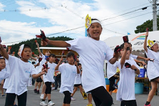 お揃いの白いTシャツを着た小学生たちが手に鳴子を持ち、そろった動きで元気に踊っている写真