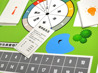 爆弾低気圧と書かれた白いカードがボード上に置かれており、その周囲には池や木のイラスト、数字のマス目、そして森・里・海・街・役場などの文字が並ぶルーレットが見える、ボードゲームの写真