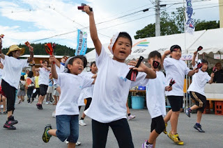 白いTシャツ姿の子どもたちが鳴子を持って元気よく踊り、先頭の男の子が大きく手を振りながらリードしている写真