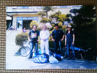 自転車や大きな荷物がそばに置かれ、4人の男女が建物の前に並んで笑顔を見せている記念写真